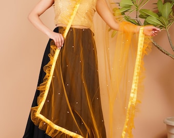 Puede incluir: Una mujer con un top corto dorado con lentejuelas, una falda negra y marrón y un dupatta amarillo transparente. La falda tiene un ribete dorado en el dobladillo. El dupatta tiene un borde con volantes y está adornado con pequeños adornos blancos.