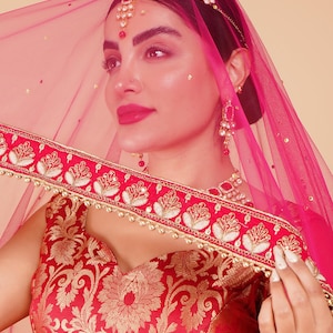 Può includere: Una donna indossa un abito tradizionale indiano. L'ensemble rosso e oro comprende un top fantasia, un copricapo decorativo e un velo rosa trasparente. L'abito è adornato con abbellimenti dorati e ricami intricati, mostrando una ricca estetica culturale.