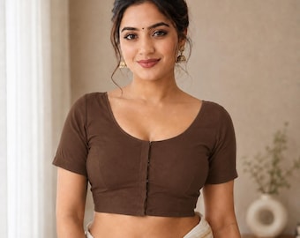 Puede incluir: Una blusa lisa de color marrón oscuro de manga corta con escote redondo y una tapeta de botones vertical en la parte delantera. La prenda es entallada con costuras visibles en el cuerpo y se combina con una tela clara drapeada en la cintura.