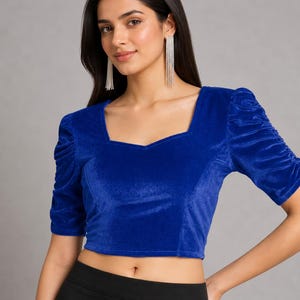 May include: A royal blue velvet crop top featuring a square neckline and short sleeves with gathered, ruched detailing. The garment is constructed from a soft, textured fabric and has a fitted silhouette that ends above the waistline.