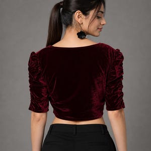 May include: A deep burgundy velvet crop top featuring a square neckline and elbow-length sleeves with gathered, ruched detailing along the shoulders and arms. The garment is paired with solid black trousers that have a visible waistband and belt loops.