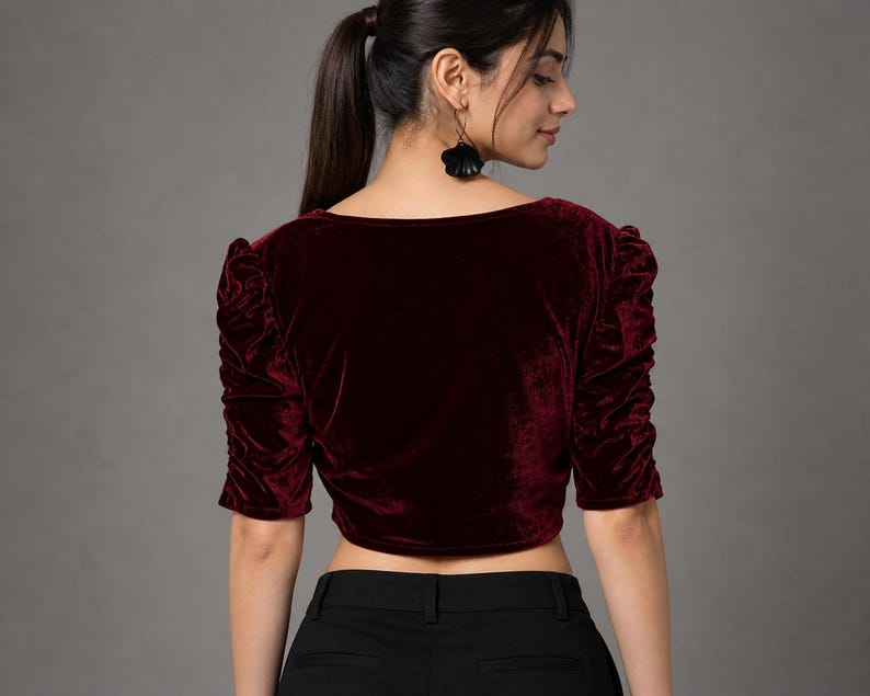 May include: A deep burgundy velvet crop top featuring a square neckline and elbow-length sleeves with gathered, ruched detailing along the shoulders and arms. The garment is paired with solid black trousers that have a visible waistband and belt loops.