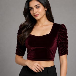 May include: A deep burgundy velvet crop top featuring a square neckline and elbow-length sleeves with gathered, ruched detailing along the shoulders and arms. The fabric has a soft, light-reflecting texture, and the garment is fitted at the waist.