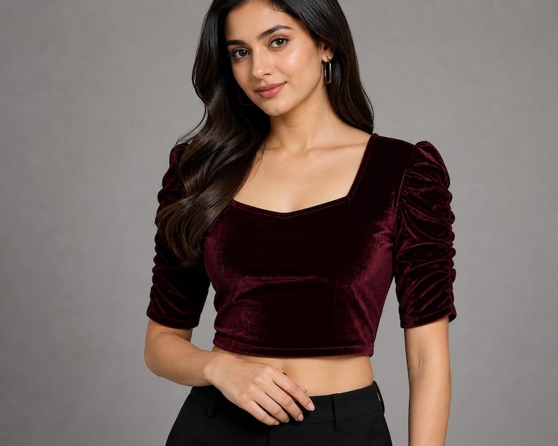 May include: A deep burgundy velvet crop top featuring a square neckline and elbow-length sleeves with gathered, ruched detailing along the shoulders and arms. The fabric has a soft, light-reflecting texture, and the garment is fitted at the waist.