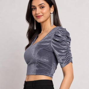 May include: A grey velvet crop top featuring a V-neckline and short sleeves with gathered, ruched detailing along the shoulders and arms. The fabric has a soft, reflective sheen characteristic of velvet, providing a textured appearance.