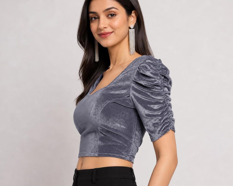 May include: A grey velvet crop top featuring a V-neckline and short sleeves with gathered, ruched detailing along the shoulders and arms. The fabric has a soft, reflective sheen characteristic of velvet, providing a textured appearance.