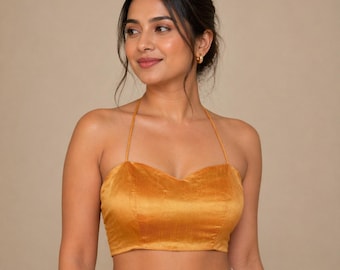 Puede incluir: Top halter mostaza con escote corazón. La parte superior está hecha de una tela suave y aterciopelada y tiene finas correas que se atan alrededor del cuello. La prenda es de estilo corto, revelando el abdomen. El fondo es de color beige neutro.