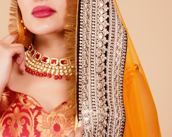 Puede incluir: Una mujer con un atuendo tradicional indio. El conjunto incluye una parte superior con estampado rojo y dorado, un collar dorado y rojo y un tocado con bordados intrincados y un borde dorado. El tocado es de color amarillo anaranjado brillante.