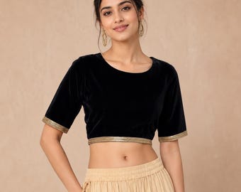 May include: Black velvet crop top with short sleeves and a gold trim along the hem and sleeve edges. The top has a round neckline. The garment is part of a two-piece outfit, paired with a beige skirt.