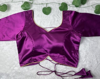 Puede incluir: Una blusa de satén púrpura vibrante con cuello cuadrado y mangas hasta el codo. La blusa presenta un frente en forma de V y un cierre trasero con lazo. El ribete dorado acentúa el escote y los bordes.