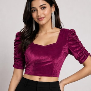 May include: A magenta velvet crop top featuring a square neckline and elbow-length puff sleeves with ruched detailing. The fabric has a soft, reflective texture, and the garment is fitted through the bodice, ending just above the waistline.