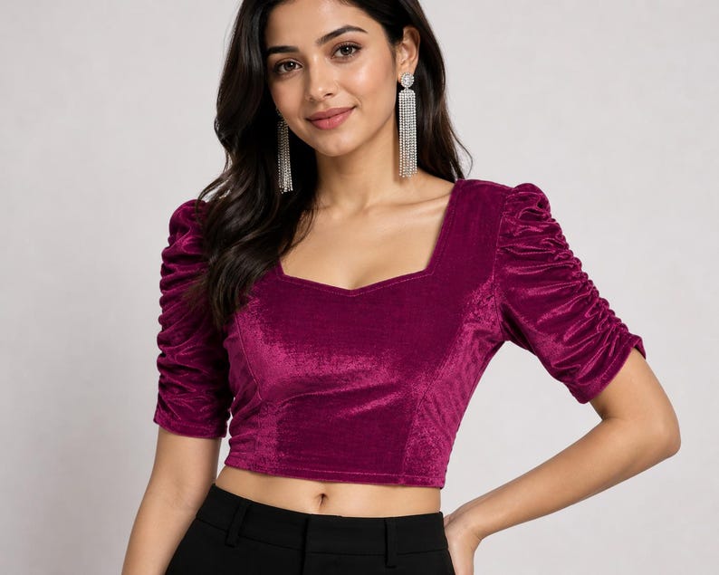 May include: A magenta velvet crop top featuring a square neckline and elbow-length puff sleeves with ruched detailing. The fabric has a soft, reflective texture, and the garment is fitted through the bodice, ending just above the waistline.