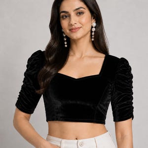 May include: A black velvet crop top featuring a square neckline and elbow-length sleeves with gathered, ruched detailing. The garment has a fitted silhouette and a smooth, dark texture, paired with light-colored bottoms visible at the waistline.