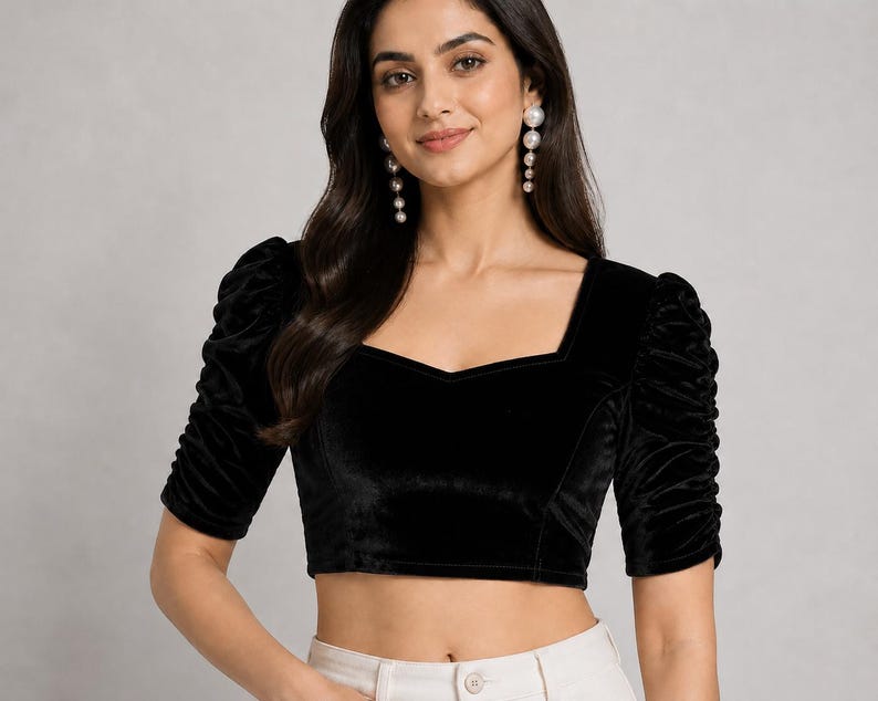 May include: A black velvet crop top featuring a square neckline and elbow-length sleeves with gathered, ruched detailing. The garment has a fitted silhouette and a smooth, dark texture, paired with light-colored bottoms visible at the waistline.