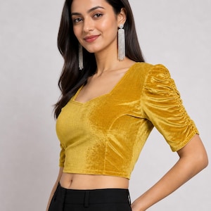 May include: A mustard yellow velvet crop top featuring a sweetheart neckline and elbow-length sleeves with gathered, ruched detailing along the outer arms. The fabric has a soft, light-reflecting texture, and the garment is paired with black trousers.