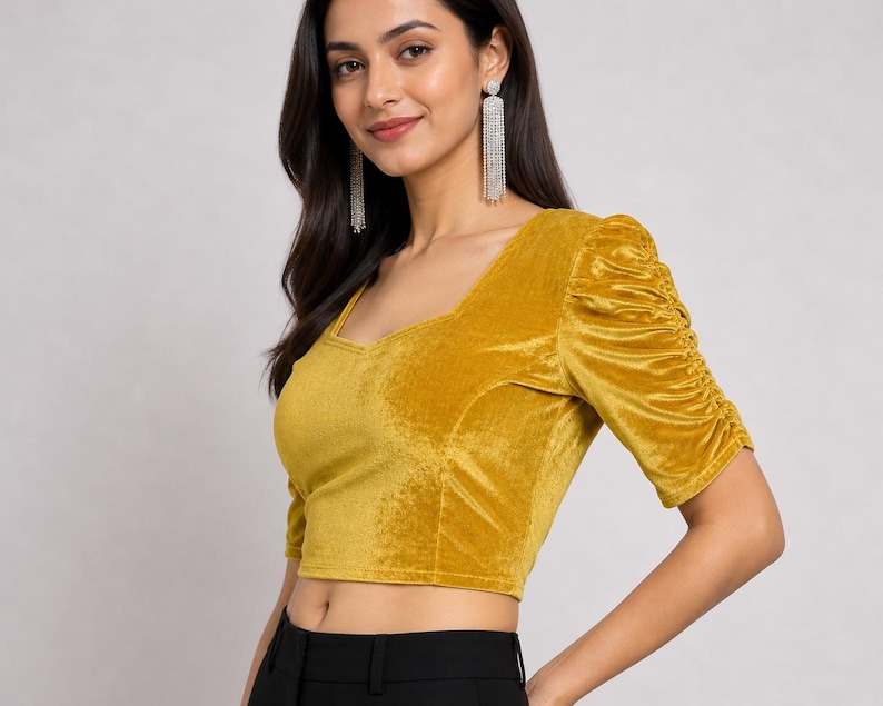 May include: A mustard yellow velvet crop top featuring a sweetheart neckline and elbow-length sleeves with gathered, ruched detailing along the outer arms. The fabric has a soft, light-reflecting texture, and the garment is paired with black trousers.