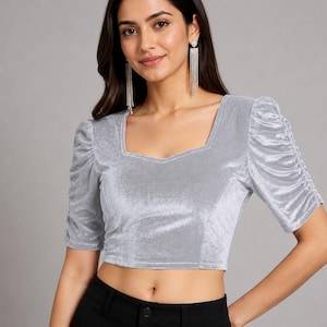 May include: A silver velvet crop top featuring a square neckline and short, puffed sleeves with gathered detailing. The fabric has a soft, reflective sheen, and the garment is fitted through the bodice with visible seam lines along the front panels.