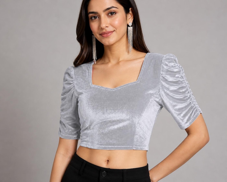 May include: A silver velvet crop top featuring a square neckline and short, puffed sleeves with gathered detailing. The fabric has a soft, reflective sheen, and the garment is fitted through the bodice with visible seam lines along the front panels.