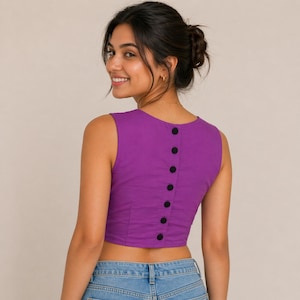 Könnte beinhalten: Ein ärmelloses, lila Crop-Top mit sechs schwarzen Knöpfen auf der Rückseite. Das Oberteil ist aus einem strukturierten Stoff gefertigt und wird mit einer blauen Jeans kombiniert. Das Model lächelt.