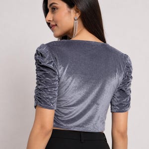 May include: A grey velvet top featuring short sleeves with gathered, ruched detailing along the shoulders and arms. The garment has a V-neckline and a cropped length, constructed from a soft, light-reflecting fabric with a smooth, plush texture.