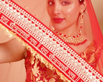 Puede incluir: Una mujer con un conjunto nupcial indio tradicional rojo y dorado. El atuendo incluye un velo rojo, un adorno para la cabeza y un collar y pendientes a juego. La tela roja tiene estampados florales dorados y texto blanco.