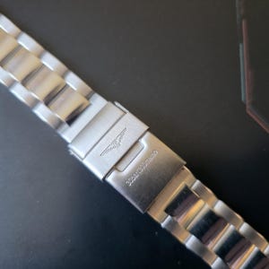 May include: A close-up of a silver watch band with a clasp. The clasp features a logo and the text "30bar/300meter". The watch band is made of interlocking metal links.