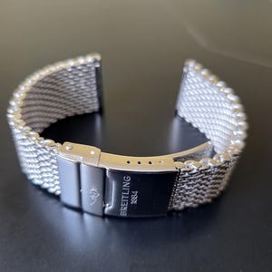 Könnte beinhalten: Ein silberfarbenes Edelstahl-Uhrenarmband mit einem Mesh-Design. Das Armband verfügt über einen Verschluss mit dem Logo "Breitling 1884". Das Armband ist für eine Uhr konzipiert und wird auf einer dunklen Oberfläche gezeigt.