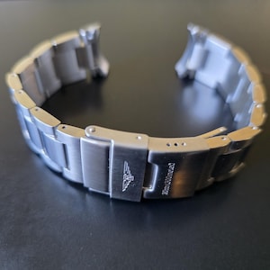 May include: A silver-toned watch band with a folding clasp. The band is made of linked metal segments. The clasp is engraved with a logo and the text "20mm/100meter".