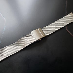 Könnte beinhalten: Ein silbernes Edelstahl-Uhrenarmband mit einem Mesh-Design. Das Armband verfügt über einen sicheren Verschluss und ist auf Langlebigkeit und Stil ausgelegt. Das Uhrenarmband wird auf einer dunklen Oberfläche gezeigt.