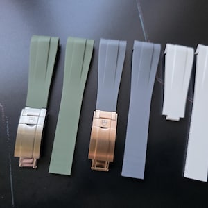 Puede incluir: Una colección de correas de reloj en varios colores, incluyendo verde oliva, gris y blanco. Las correas tienen cierres metálicos en tonos dorados y oro rosa. Las correas de reloj se muestran sobre una superficie oscura.