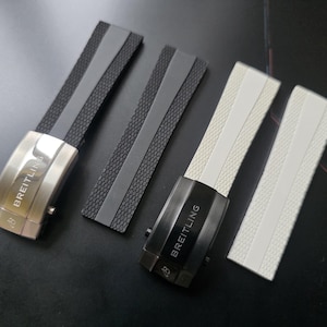 May include: Four watch straps with metal clasps. Two are black with grey accents, and two are white with white accents. The clasps are silver and black, each with the brand name "BREITLING" engraved.