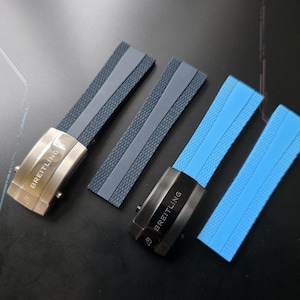 Puede incluir: Cuatro correas de reloj con cierres metálicos. Dos correas son azul marino con cierres plateados, y dos son azul claro con cierres negros. Los cierres están grabados con la palabra "BREITLING". Las correas tienen una superficie texturizada.