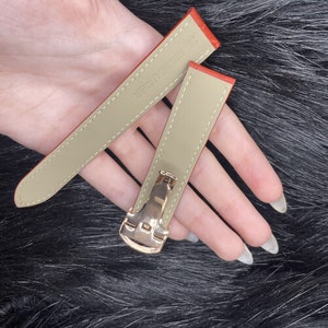 Puede incluir: Dos correas de reloj de cuero naranja con hebillas doradas. Las correas son de color beige en el interior y tienen el texto "Genuine Leather" impreso en una de las correas.