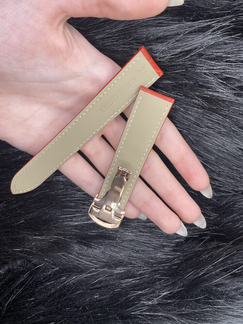 Puede incluir: Dos correas de reloj de cuero naranja con hebillas doradas. Las correas son de color beige en el interior y tienen el texto "Genuine Leather" impreso en una de las correas.