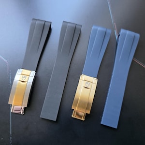 Puede incluir: Cuatro correas de reloj en negro y gris azulado. Dos correas tienen cierres dorados y plateados, y dos tienen cierres dorados. Las correas están hechas de un material flexible y los cierres tienen un logotipo. Las correas son para un reloj.