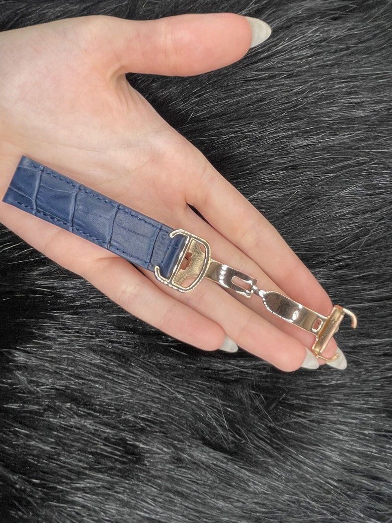 Puede incluir: Una correa de reloj de cuero azul con hebilla y cierre dorados.