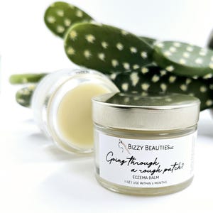 May include: A small glass jar with a silver lid containing a white cream. The jar has a white label with the text "Bizzy Beauties LLC Going through a rough patch? ECZEMA BALM 1 OZ | USE WITHIN 6 MONTHS".