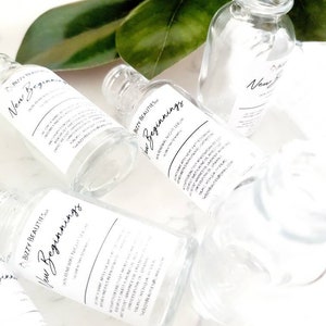 May include: Three clear glass bottles with labels that read "Bizzy Beauties New Beginnings Overnight Renewal Night Serum". The bottles are on a white surface with green leaves in the background.