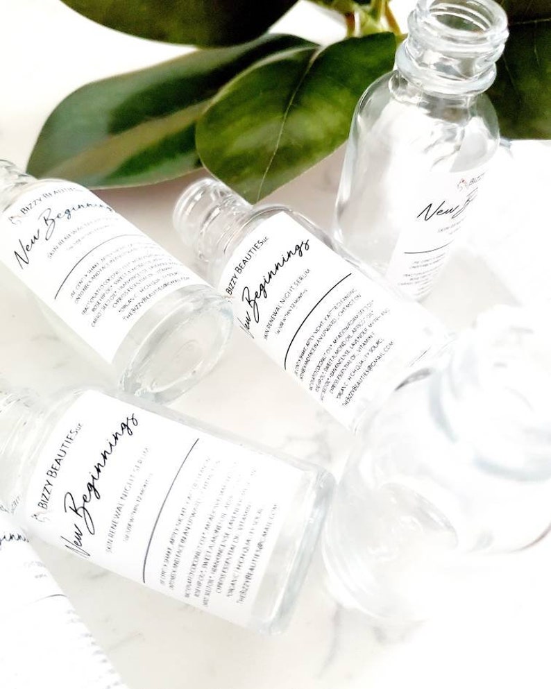 May include: Three clear glass bottles with labels that read "Bizzy Beauties New Beginnings Overnight Renewal Night Serum". The bottles are on a white surface with green leaves in the background.
