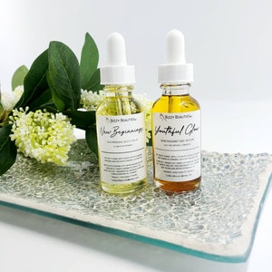May include: Two amber glass bottles with dropper tops, labeled "Bizzy Beauties" and containing facial serums. The bottles are labeled "New Beginnings Skin Renewal Night Serum" and "Youthful Glow Skin Radiant Day Serum".