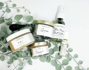 May include: Four glass jars with gold lids containing skincare products. The jars are labeled "Green", "3 in 1", "Reurnd", and "Blue Tangy Plant Extract". The jars are arranged on a white surface with green leafy branches.