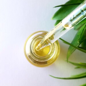 May include: A clear glass bottle with a dropper filled with a yellow liquid. The dropper is inserted into the bottle and the tip is submerged in the liquid. The bottle is on a white surface with green leaves in the background.