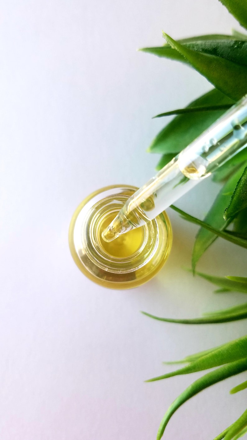 May include: A clear glass bottle with a dropper filled with a yellow liquid. The dropper is inserted into the bottle and the tip is submerged in the liquid. The bottle is on a white surface with green leaves in the background.