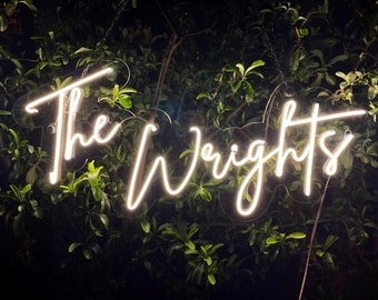 Pode incluir: Um letreiro de néon branco com o texto "The Wrights" pendurado em um galho de árvore. O letreiro está iluminado contra um fundo escuro de folhagem verde.