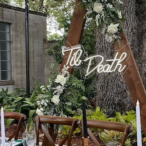 Può includere: Una mise en place per un ricevimento di nozze con una tovaglia verde, candele bianche e un centrotavola floreale. La tavola è apparecchiata con piatti bianchi, tovaglioli verdi e posate dorate. Un cartello al neon con la scritta "Til Death" è visibile sullo sfondo.