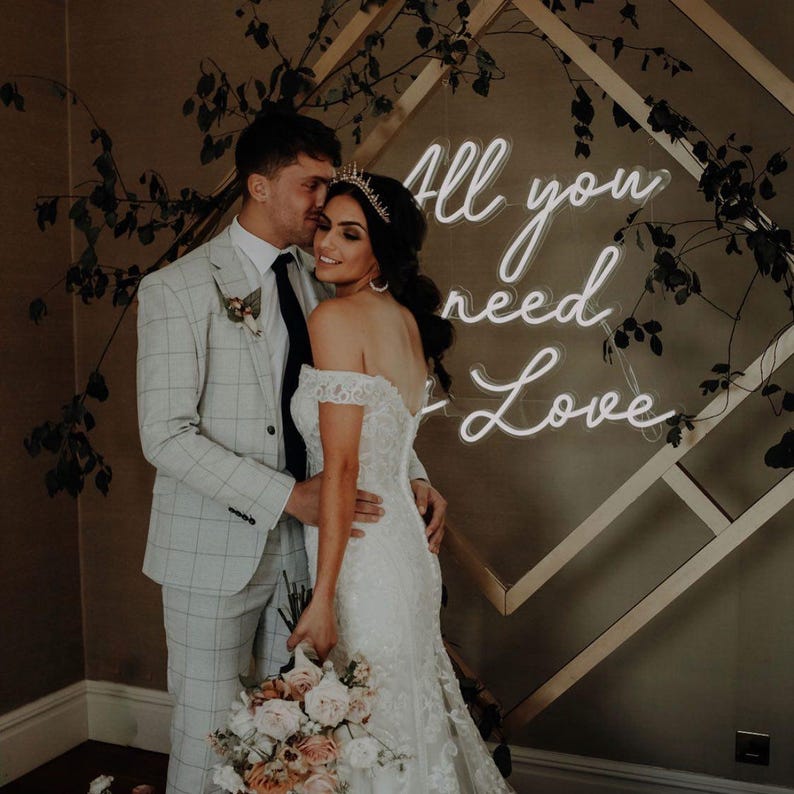 K&ouml;nnte beinhalten: Ein Paar umarmt sich vor einer Wand, die mit einem Neonzeichen mit der Aufschrift "All you need is Love" geschm&uuml;ckt ist. Die Braut tr&auml;gt ein wei&szlig;es schulterfreies Brautkleid aus Spitze und ein Diadem. Der Br&auml;utigam tr&auml;gt einen hellgrauen Anzug mit Karomuster. Die Braut h&auml;lt einen Blumenstrau&szlig;.