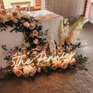K&ouml;nnte beinhalten: Ein Hochzeitstisch, dekoriert mit rosa und wei&szlig;en Rosen, Gr&uuml;nzeug und einem Neon-Schild mit der Aufschrift "The Pennies". Der Tisch ist mit einer wei&szlig;en Tischdecke bedeckt.