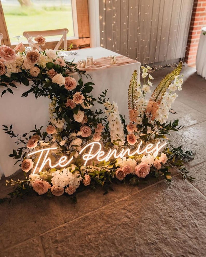K&ouml;nnte beinhalten: Ein Hochzeitstisch, dekoriert mit rosa und wei&szlig;en Rosen, Gr&uuml;nzeug und einem Neon-Schild mit der Aufschrift "The Pennies". Der Tisch ist mit einer wei&szlig;en Tischdecke bedeckt.