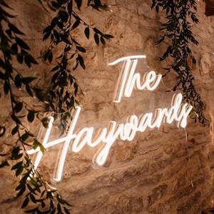 K&ouml;nnte beinhalten: Ein Neon-Schild mit der Aufschrift "The Haywards" in geschwungener Schrift, montiert an einer strukturierten Steinmauer. Gr&uuml;ne Bl&auml;tter und kleine Lichter umrahmen das Schild und schaffen eine warme, einladende Atmosph&auml;re. Ideal f&uuml;r Hochzeiten oder Veranstaltungen.