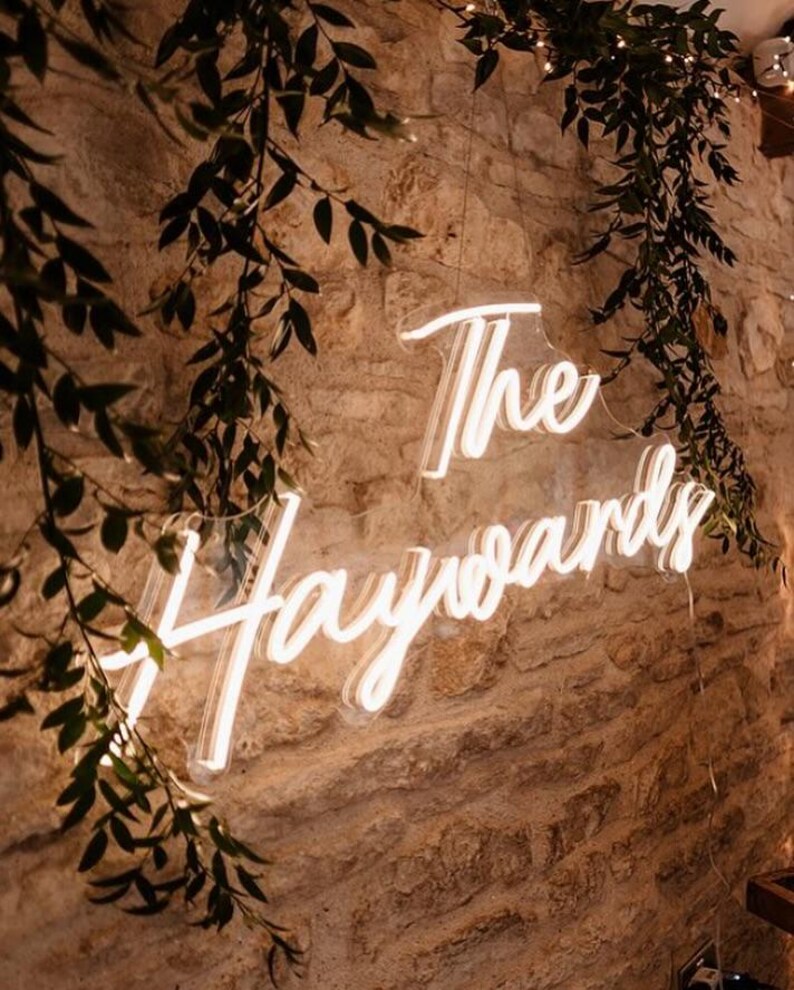 K&ouml;nnte beinhalten: Ein Neon-Schild mit der Aufschrift "The Haywards" in geschwungener Schrift, montiert an einer strukturierten Steinmauer. Gr&uuml;ne Bl&auml;tter und kleine Lichter umrahmen das Schild und schaffen eine warme, einladende Atmosph&auml;re. Ideal f&uuml;r Hochzeiten oder Veranstaltungen.